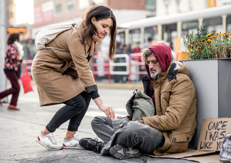 Young Woman Giving Money To Homeless Beggar Man Sitting in City. Stock