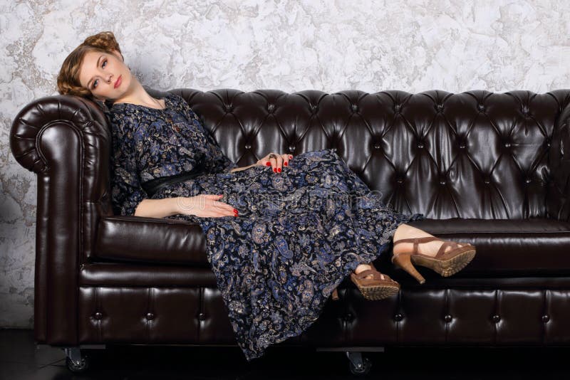 Young woman with hairdo in dress lies on black leather sofa