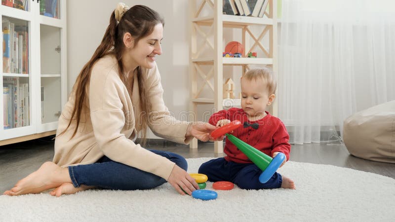 Young woman helping her baby son building toy tower with colorful rings. Baby development, child playing games, education and