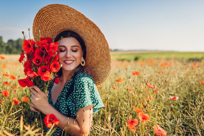 Young woman holding bouquet of poppies flowers walking in summer field. Stylish happy girl wearing straw hat