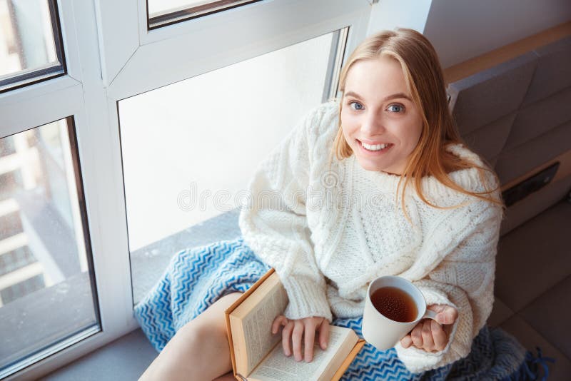 Young woman at home sitting on window sill winter concept drinking tea reading book