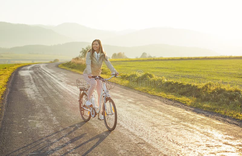 Young woman in jacket rides a bicycle over country road, afternoon sun shines at background