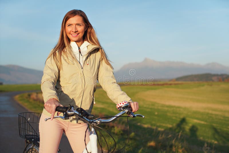 Young woman in jacket standing with a bicycle on asphalt country road, afternoon sun shines on fields meadows and mountains