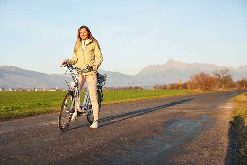 Young woman in jacket standing with a bicycle over country road, afternoon sun shines at mount Krivan peak - Slovak symbol -