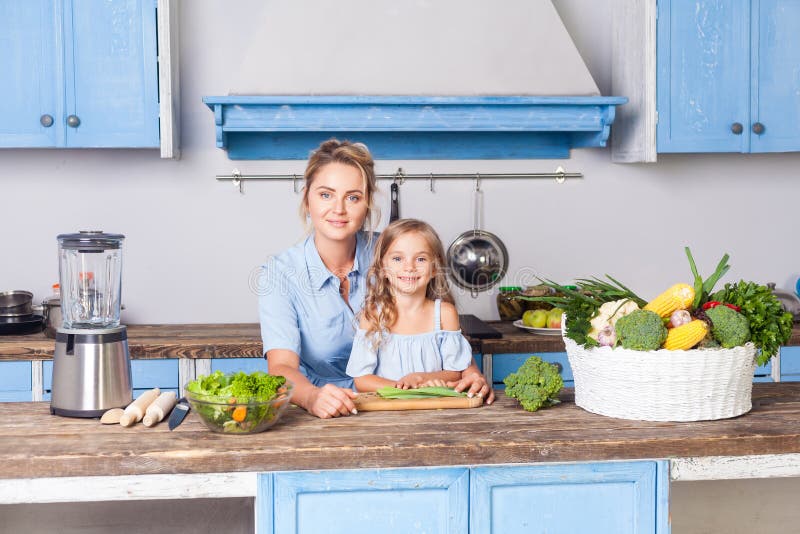 Young woman and lovely girl sitting in modern kitchen, looking at camera, mother and daughter cooking salad together stock photography Young woman and lovely girl sitting in modern kitchen, looking at camera, mother and daughter cooking salad together stock photography