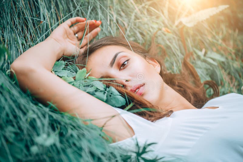 Young woman lying on the grass. Beauty woman lying on the field and looking on camera. Beautiful brunette Girl lying on the meadow