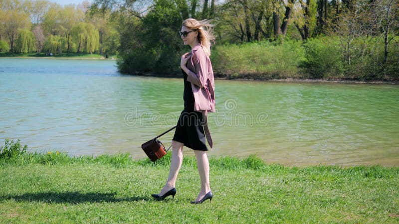 A young woman in office clothes walks in the park on the grass along the lake contentedly with a handbag in her hand and