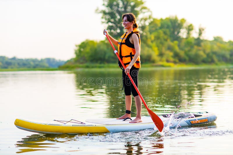Young Woman in Orange Life Jacket on Supboard at River Stock Photo