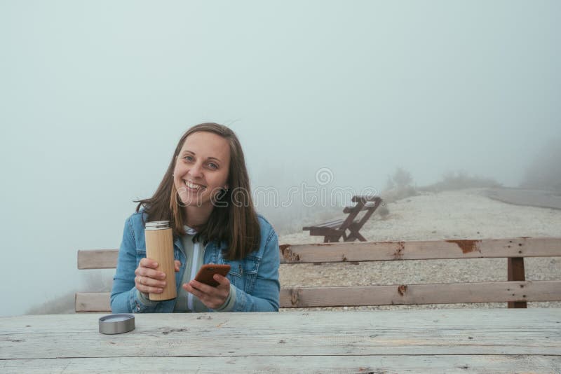 Young woman with phone sitting on Wooden bench and drinking hot