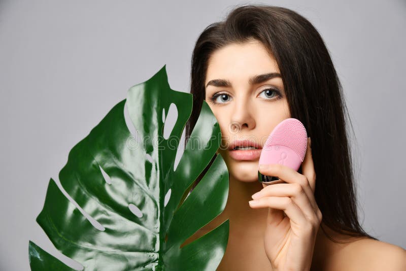 Young woman with pink face exfoliator brush silicone cleansing device for sensitive normal skin with tropical palm leaf on gray stock photography Young woman with pink face exfoliator brush silicone cleansing device for sensitive normal skin with tropical palm leaf on gray stock photography