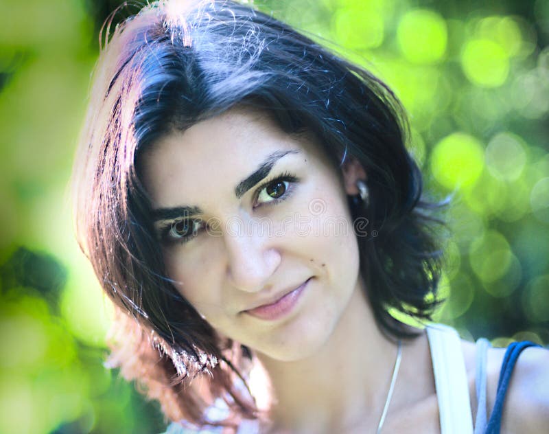 Young woman portrait at the green bokeh background. Woman is brunette, background is number of trees.