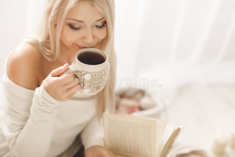 Young beautiful woman - blonde, wearing a warm knit short dress handmade, light - beige, reading a book with a cup of coffee in his hand lying on the couch at home in the bedroom. Young beautiful woman - blonde, wearing a warm knit short dress handmade, light - beige, reading a book with a cup of coffee in his hand lying on the couch at home in the bedroom.