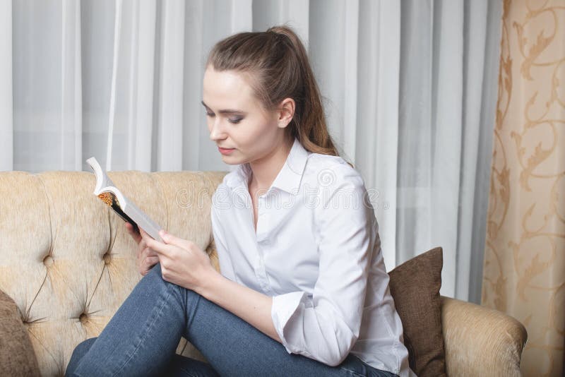 Hübsche junge Frau sitzt auf sofa und liest ein Buch.