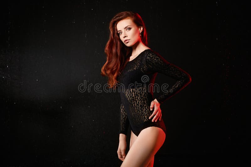 Young woman with red hair and perfect body in black bodysuit posing at dark background. Beautiful redhaired model girl
