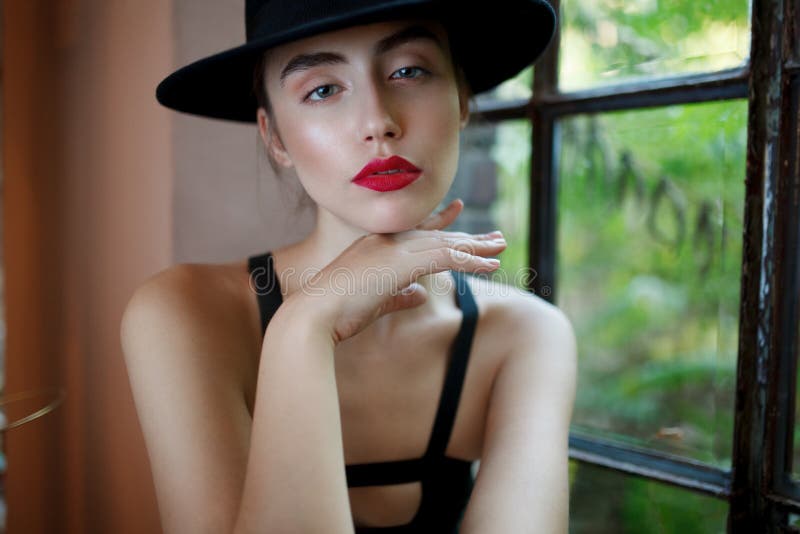 Young woman with red lips, in black hat, sitting on chair, touching her chin delicate with hand, behind a window. stock photo Young woman with red lips, in black hat, sitting on chair, touching her chin delicate with hand, behind a window. stock photo