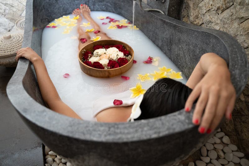 Young woman relaxing in black stone bath with tropical flowers and rose petals. Skin treatment, luxury spa concept