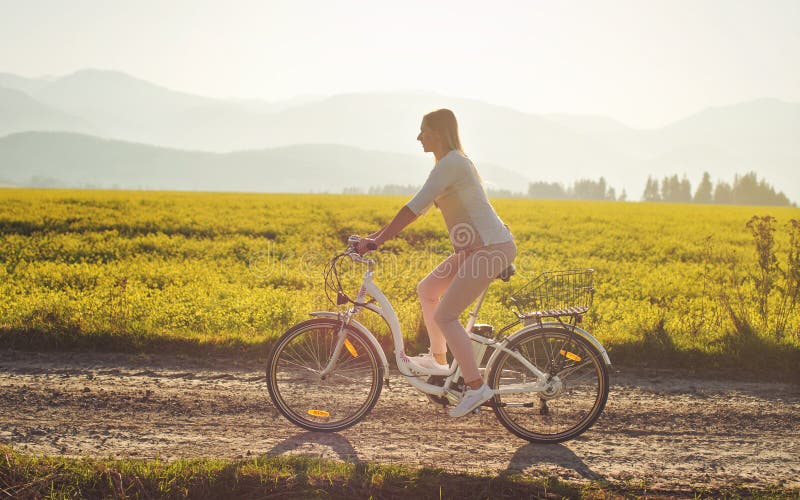 Young woman rides electric bike over dusty country road, strong afternoon sun backlight in background shines on yellow flowers. Field, view from side