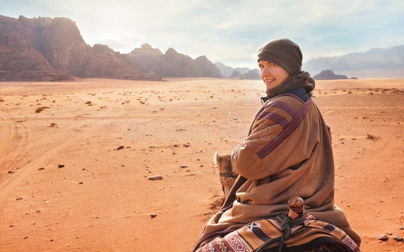 Young woman riding camel in desert, looking back over her shoulder, smiling. It`s quite cold so she is wearing traditional Bedoui