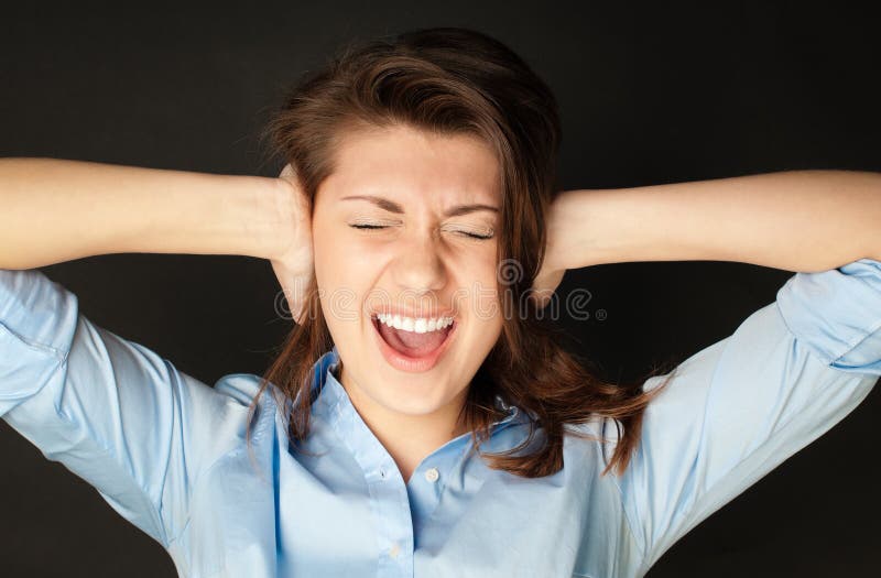 Beautiful young woman in blue shirt with dark hair screaming. Hands at her ears, mouth wide open, eyes closed, dark background. Beautiful young woman in blue shirt with dark hair screaming. Hands at her ears, mouth wide open, eyes closed, dark background.