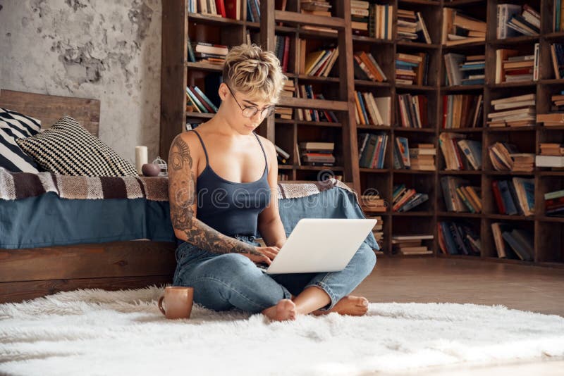Side Hustle. Young woman short hair in glasses sitting on floor working online on laptop concentrated with cup of coffee