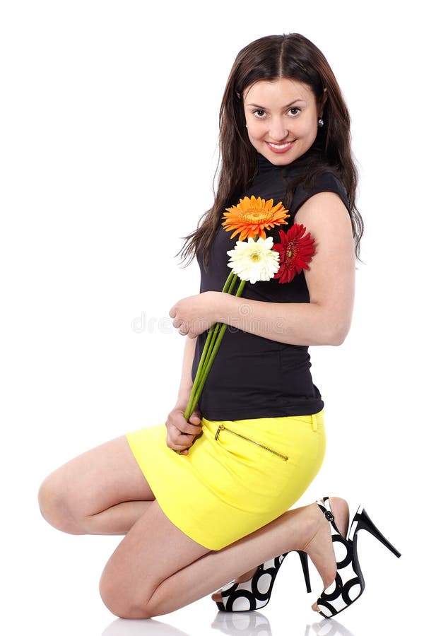 Young woman in short yellow skirt with gerberas squats in studio on white background