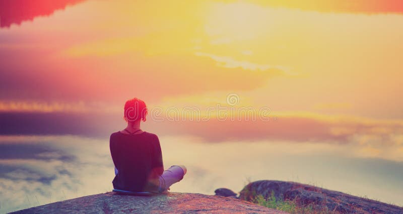 Young woman sitting enjoying peaceful moment of beautiful colorful sunset. In the reflection of lake water sees clouds