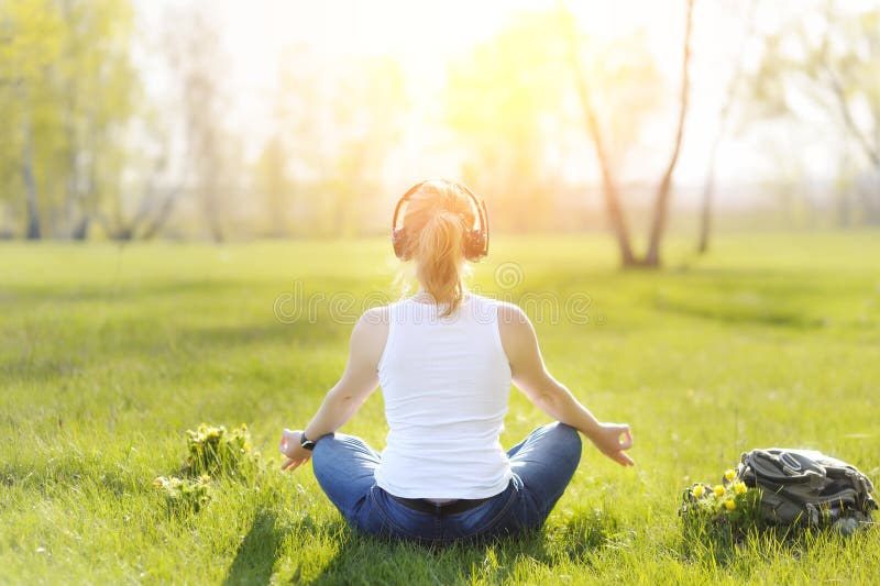 Young woman sitting on grass in Park and meditating and listening to music on headphones.