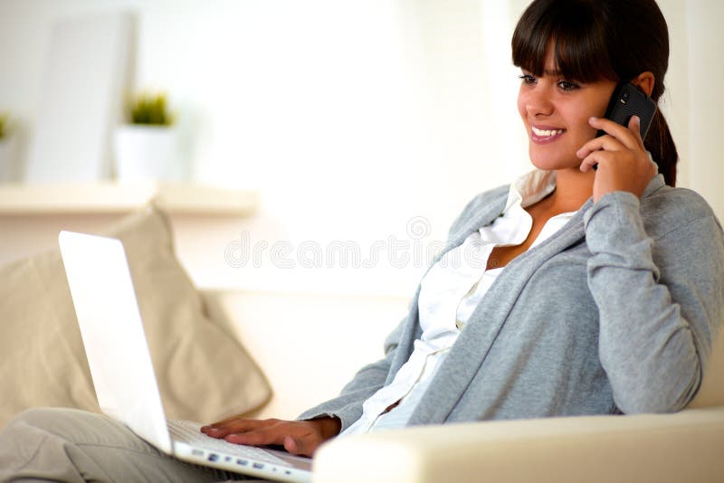 Portrait of a young woman sitting on sofa in front of her laptop at home indoor speaking on cellphone. Portrait of a young woman sitting on sofa in front of her laptop at home indoor speaking on cellphone