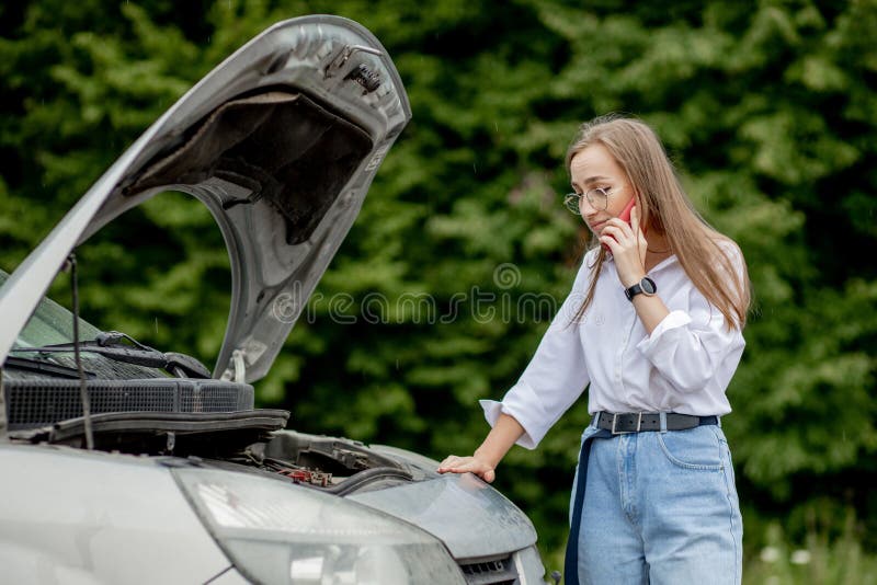 Young woman standing near broken down car with popped up hood having trouble with her vehicle. Waiting for help tow truck or