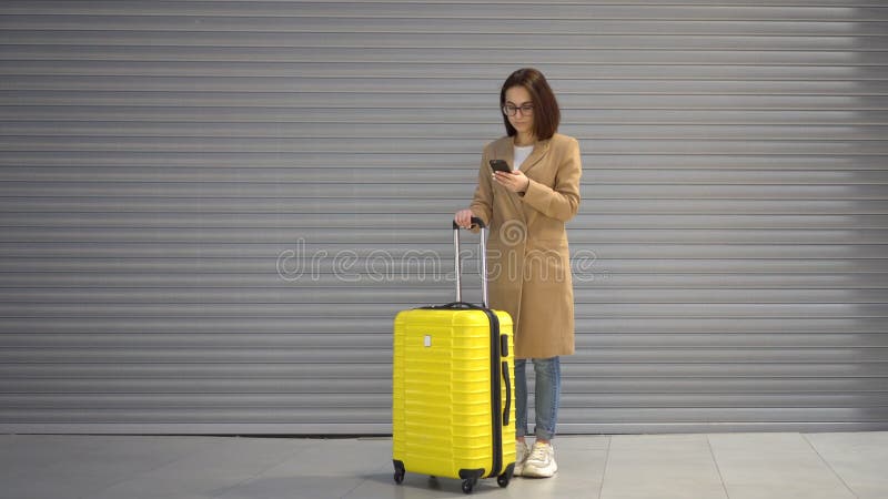 A young woman stands with a suitcase and a phone in her hands. Girl in a coat on a gray background.