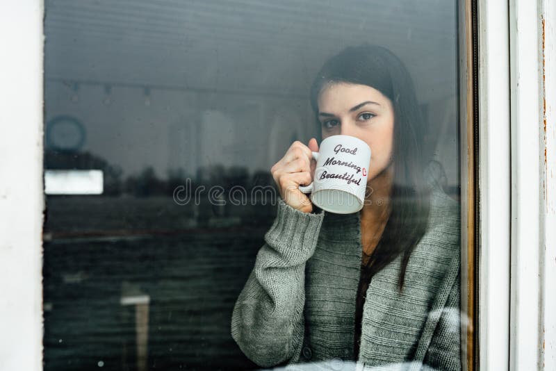 Young woman staying at home drinking coffee/tea,looking trough the window.Starting the day,morning ritual.Quaratine self isolation