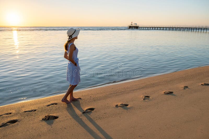 Young woman in straw hat and a dress walking alone on empty sand beach at sunset sea shore. Lonely girl looking at horizon over