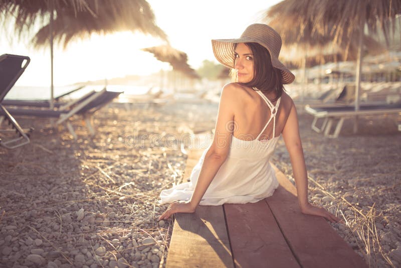 Young woman in straw hat sitting on a tropical beach, enjoying sand and sunset. Laying in the shade of palm tree parasols