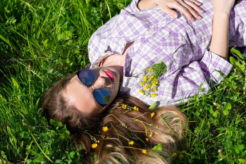 Young woman in sunglasses and little yellow flowers in blonde hair and shirt pocket is lying down and resting on green sunlit gras