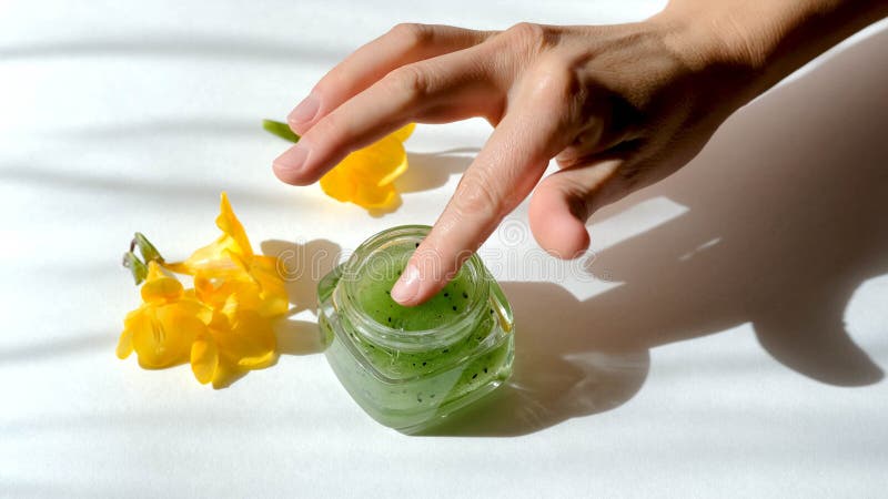 Young woman takes natural green kiwi fruits scrub from glass jar. Pure sugar scrub, peeling, face mask, girls hand and yellow freesia flowers in morning sun rays. Girl doing skincare procedure. Young woman takes natural green kiwi fruits scrub from glass jar. Pure sugar scrub, peeling, face mask, girls hand and yellow freesia flowers in morning sun rays. Girl doing skincare procedure