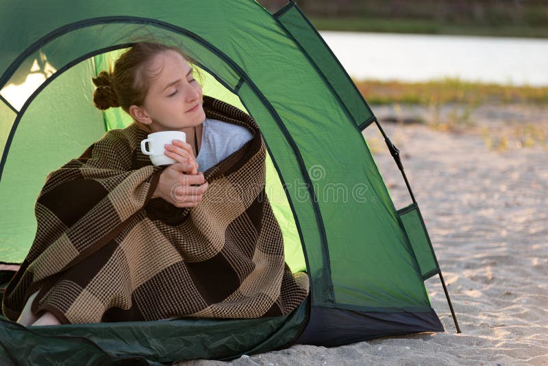 Young woman in tent drinks morning tea. Camping holiday in countryside