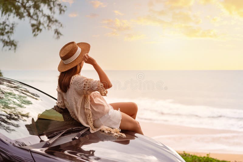 Young woman traveler sitting on a car and looking a beautiful sunset at the beach while travel driving road trip