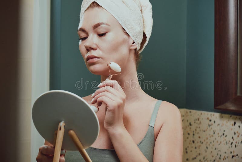 Young Woman Using Jade Facial Roller For Face Massage Sitting In Bathroom Looking In The Mirror