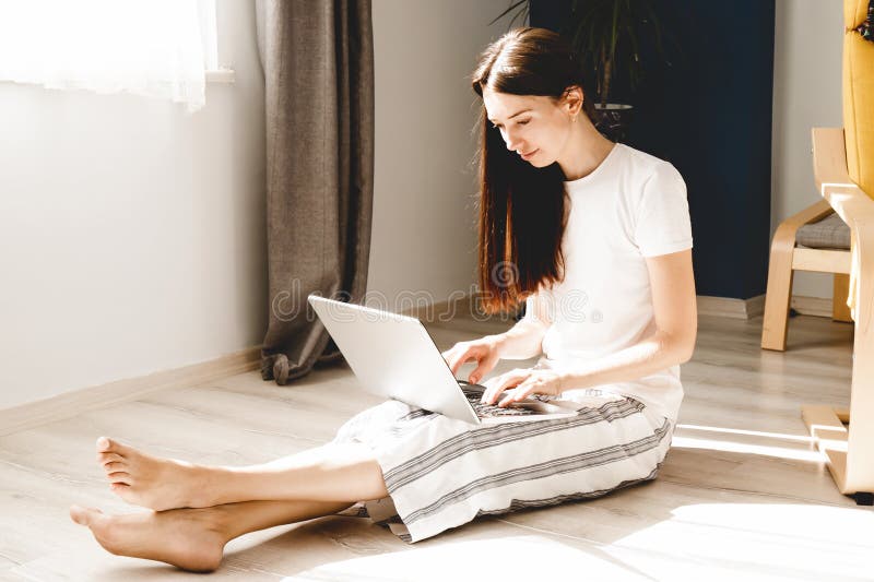 Young woman using laptop at home for remote work. Woman sitting on the floor on sunny day stock photo