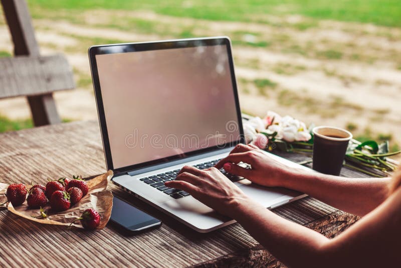 Young woman using and typing laptop computer at rough wooden table with coffee cup, strawberries, bouquet of peonies flowers