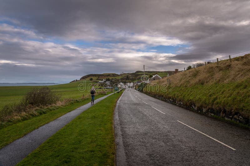 Young woman walking lonely along a road towards to a coastal city on a cold and rainy day with fields and the sea aside while