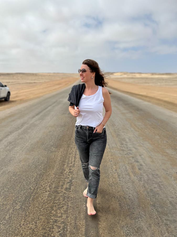 Young woman walks on highway in desert. Travel Africa, Namibia. Girl on center of the road, barefoot. Skeleton coast asphalt. Young woman walks on highway in desert. Travel Africa, Namibia. Girl on center of the road, barefoot. Skeleton coast asphalt.