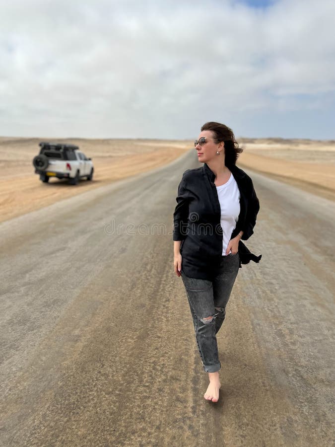 Young woman walks on highway in desert. Travel Africa, Namibia. Girl on center of the road, barefoot. Skeleton coast asphalt. Young woman walks on highway in desert. Travel Africa, Namibia. Girl on center of the road, barefoot. Skeleton coast asphalt.