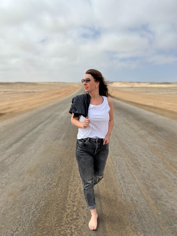 Young woman walks on highway in desert. Travel Africa, Namibia. Girl on center of the road, barefoot. Skeleton coast asphalt. Young woman walks on highway in desert. Travel Africa, Namibia. Girl on center of the road, barefoot. Skeleton coast asphalt.