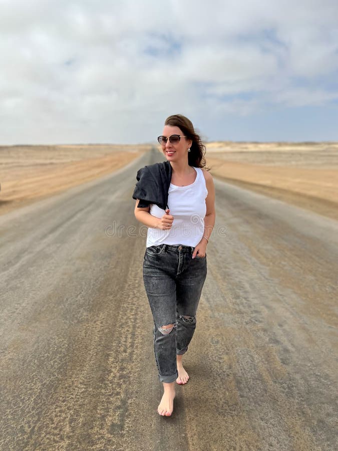 Young woman walks on highway in desert. Travel Africa, Namibia. Girl on center of the road, barefoot. Skeleton coast asphalt. Young woman walks on highway in desert. Travel Africa, Namibia. Girl on center of the road, barefoot. Skeleton coast asphalt.