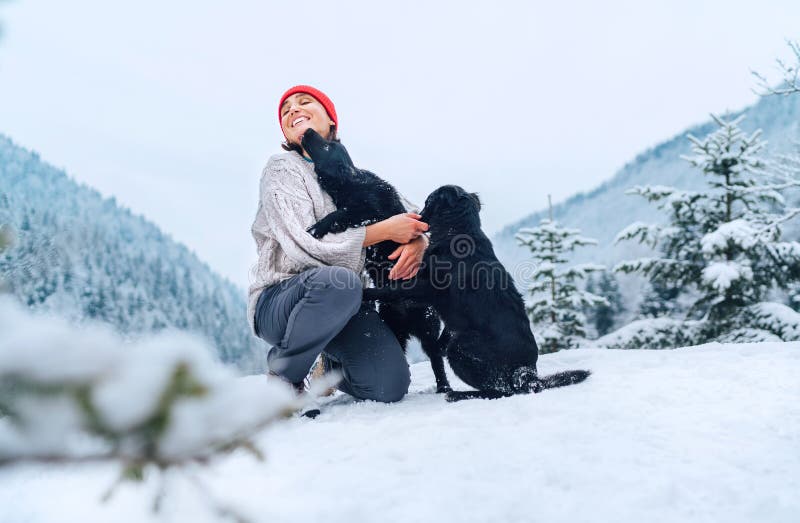 A young woman in warm clothes walking her 2 dogs in a picturesque snowy mountain outdoor. Female laughing and playing with pets