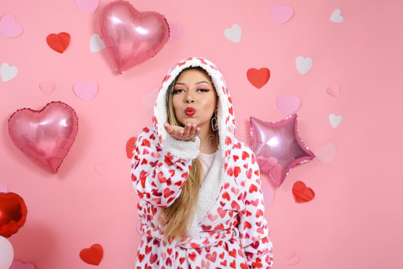 Young woman wearing bathrobe with red hearts blowing a kiss. Valentine`s day background with balloons and hearts