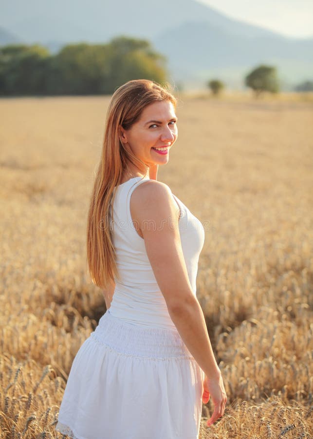 Young woman in white dress looking back over her shoulder, smiling, afternoon sun lit wheat field behind her stock image Young woman in white dress looking back over her shoulder, smiling, afternoon sun lit wheat field behind her stock image