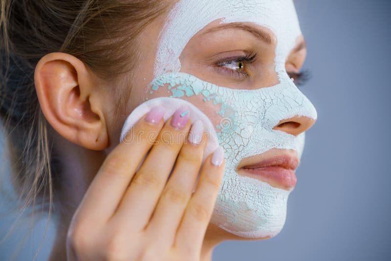 Young woman with white dried mud mask on face being removing cosmetic with cotton swab. Teen girl taking care of oily skin. Beauty treatment. Skincare. Young woman with white dried mud mask on face being removing cosmetic with cotton swab. Teen girl taking care of oily skin. Beauty treatment. Skincare