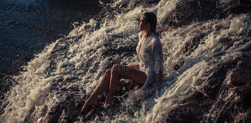 Young woman in white shirt and bikini sits on rock in water flow